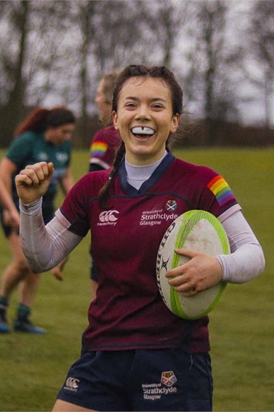 Strathclyde Women's Rugby player in field, holding rugby ball and smiling to the camera in celebration.