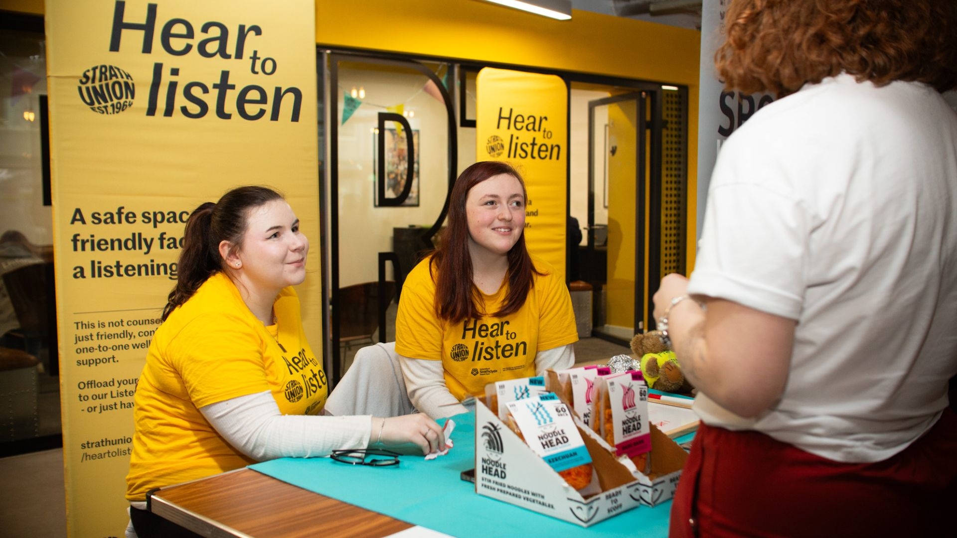 Two Hear to Listen volunteers sitting behind a stall, smiling at a student who is talking to them.