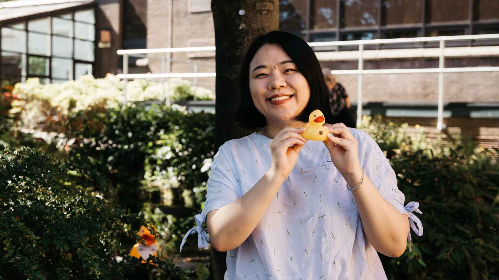 Person on Strathclyde campus, holding a small yellow rubber duck and smiling at the camera.