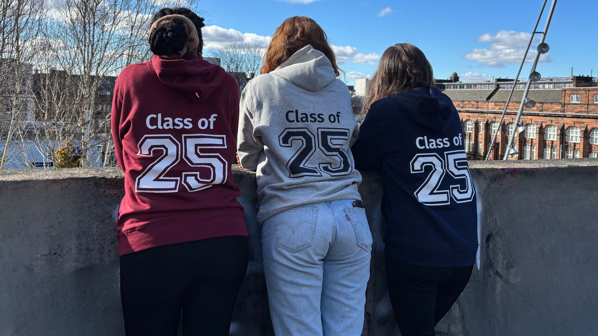 Students wearing graduation hoodies looking out over Rottenrow Gardens over a wall at the top