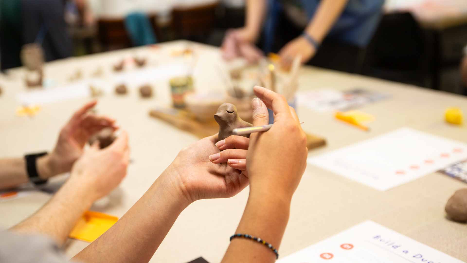 Close-up of someone sculpting a duck out of clay, other people sculpting with clay in the background.