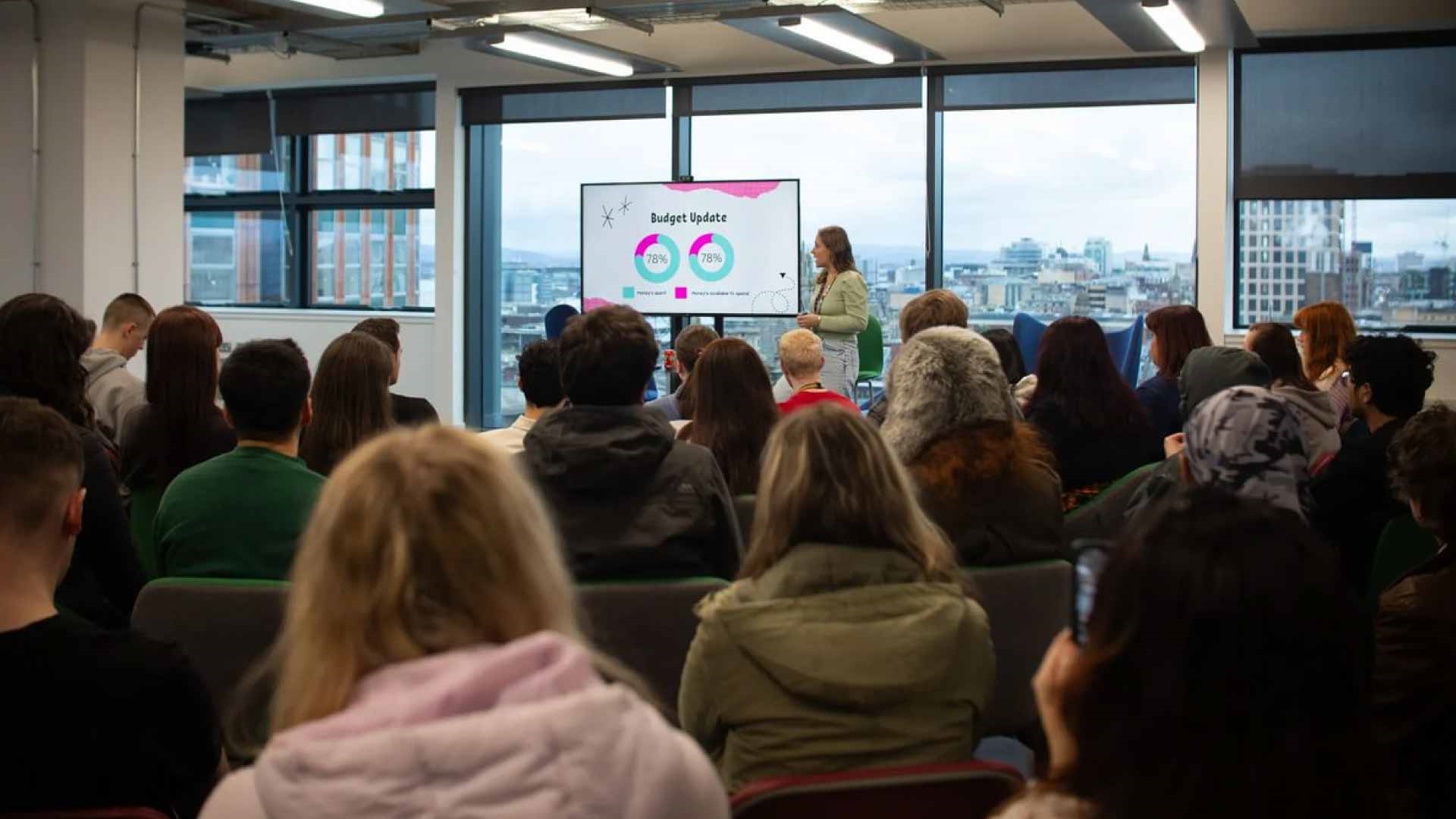 Person pointing to a screen, giving a speech to a crowd of students, with Glasgow visible in the background through a window.