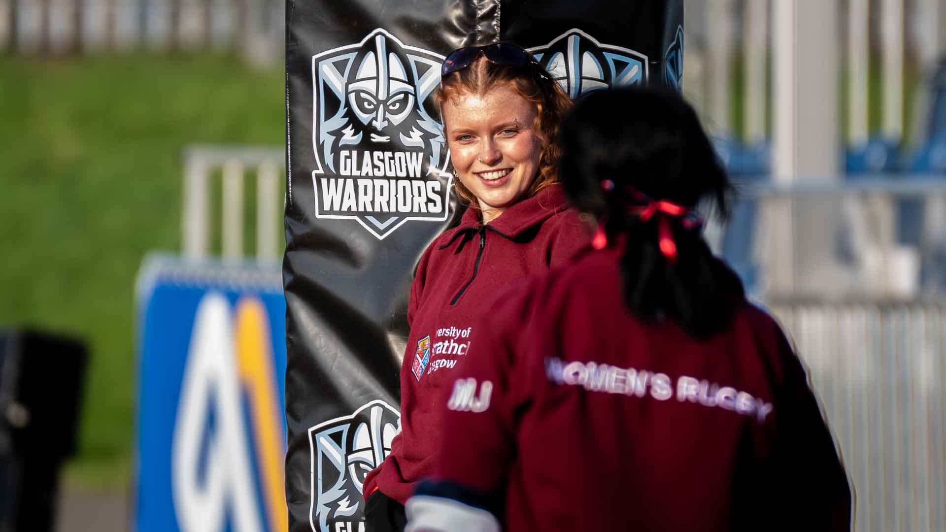 Strathclyde Women's Rugby players in field, one smiling, with Glasgow Warriors logo visible in the background.