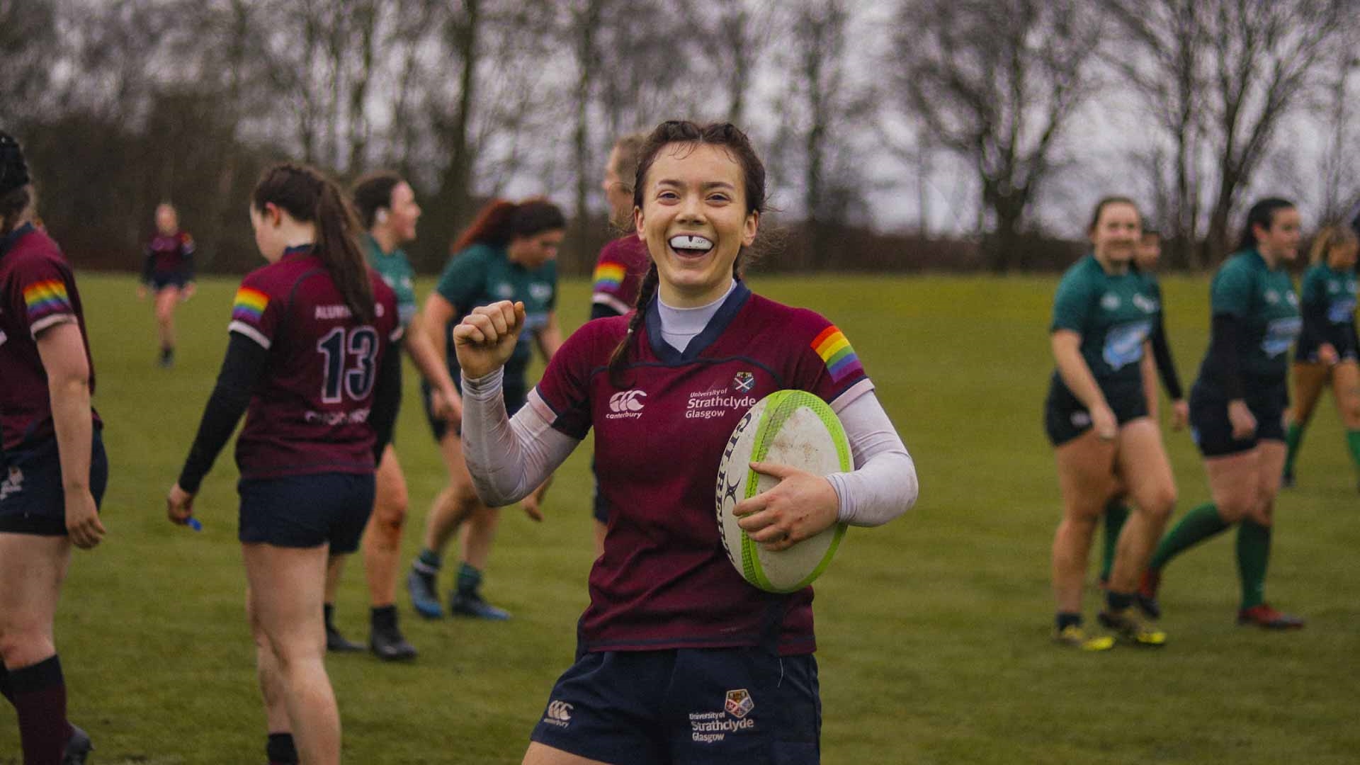 Strathclyde Women's Rugby player in field, holding rugby ball and smiling to the camera in celebration.