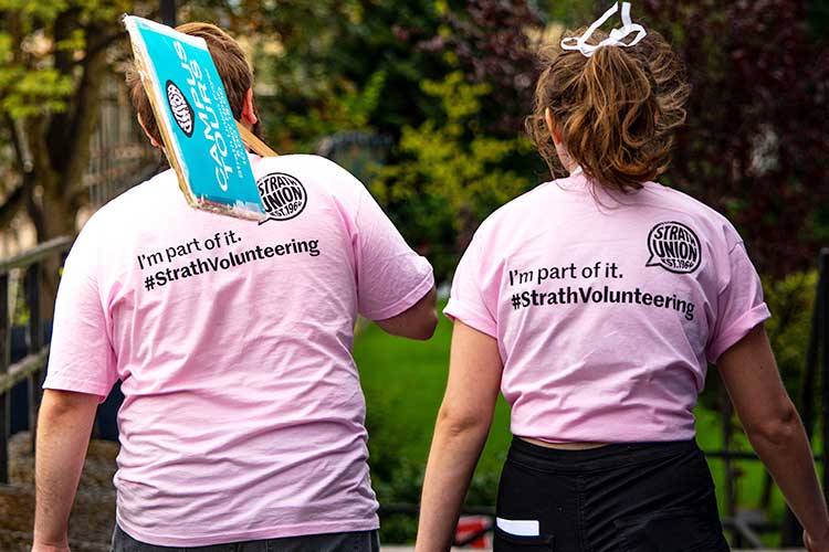 Two students walking away from the camera wearing pink t-shirts that read 'I'm part of it. #StrathVolunteering' across the back
