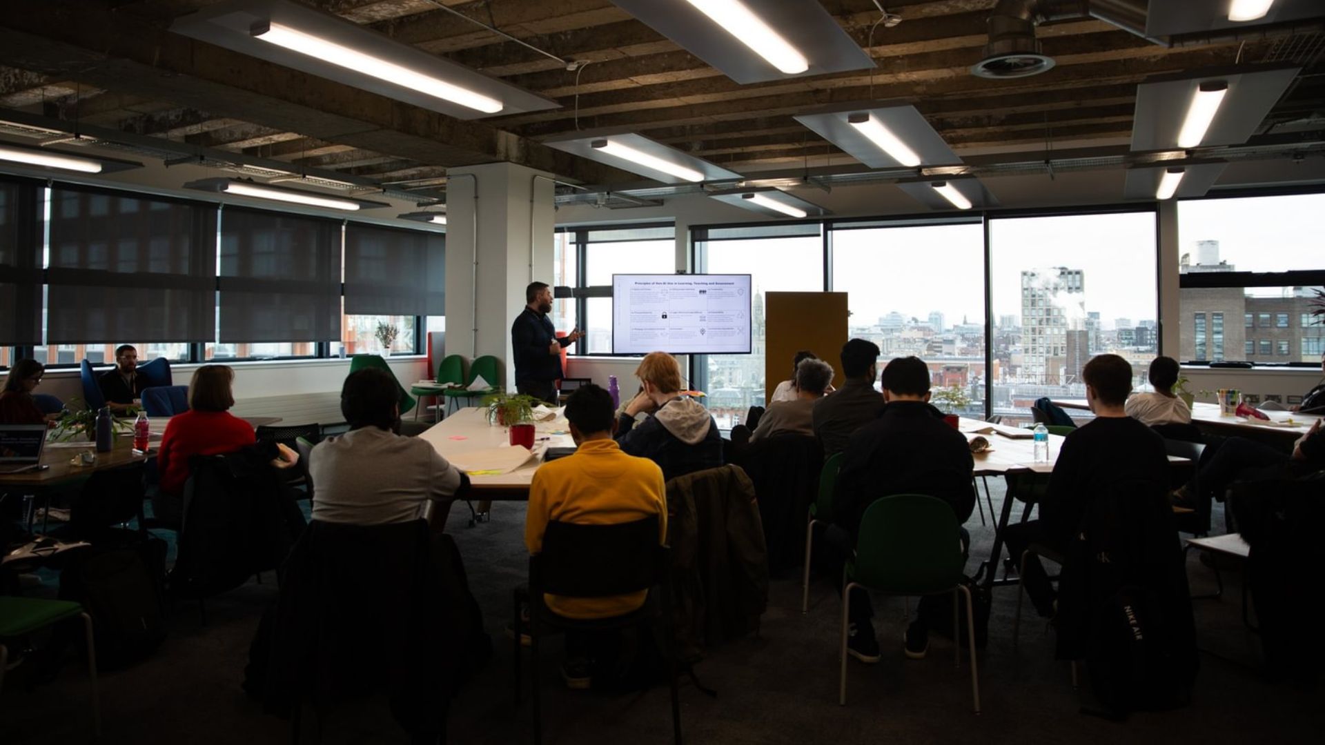 Person pointing to a screen, giving a speech to a crowd of students, with Glasgow visible in the background through a window.