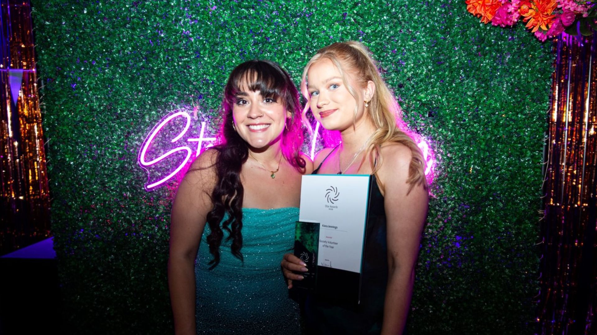 Student volunteer and Exec Officer standing in front of Strath Union flower wall, holding a STAR award and smiling at the camera.