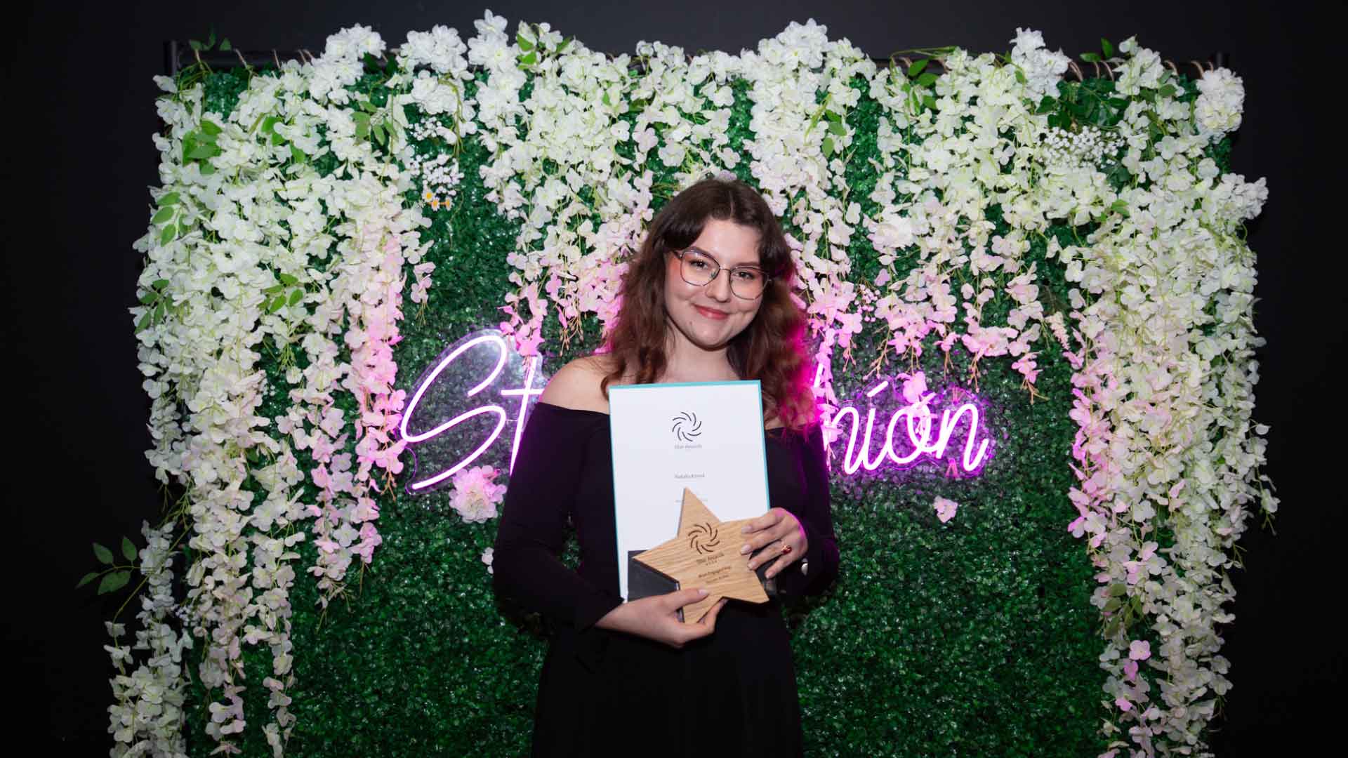 Students volunteer standing in front of Strath Union flower wall, holding their award and smiling at the camera
