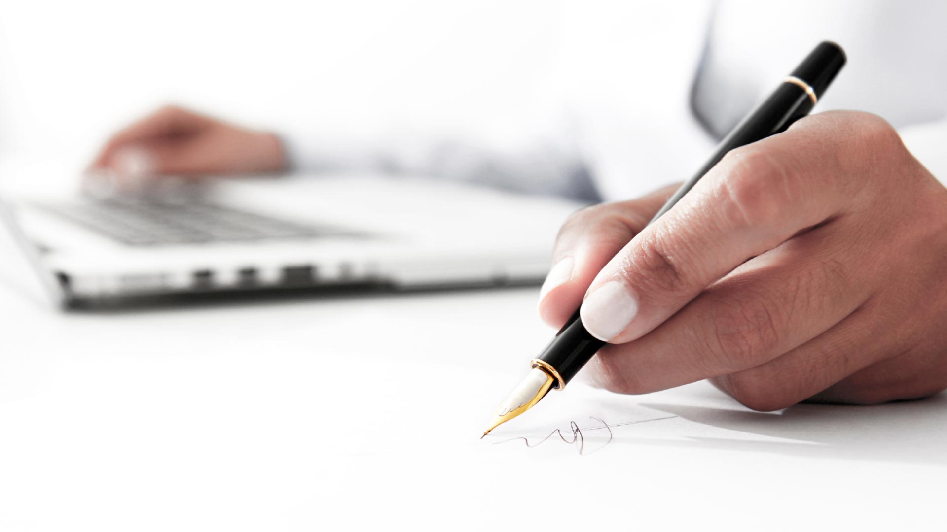 Closeup of a person's hands, using a laptop and taking a note with a pen.
