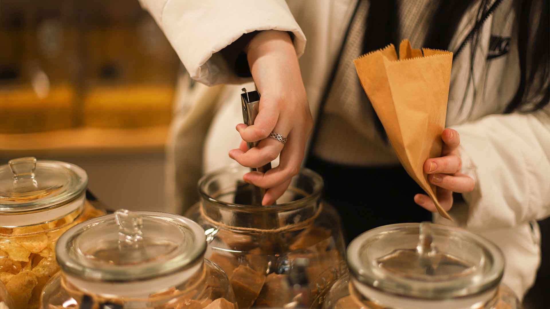 Someone holding a brown paper bag and reaching into a large biscotti jar with a pair of tongs