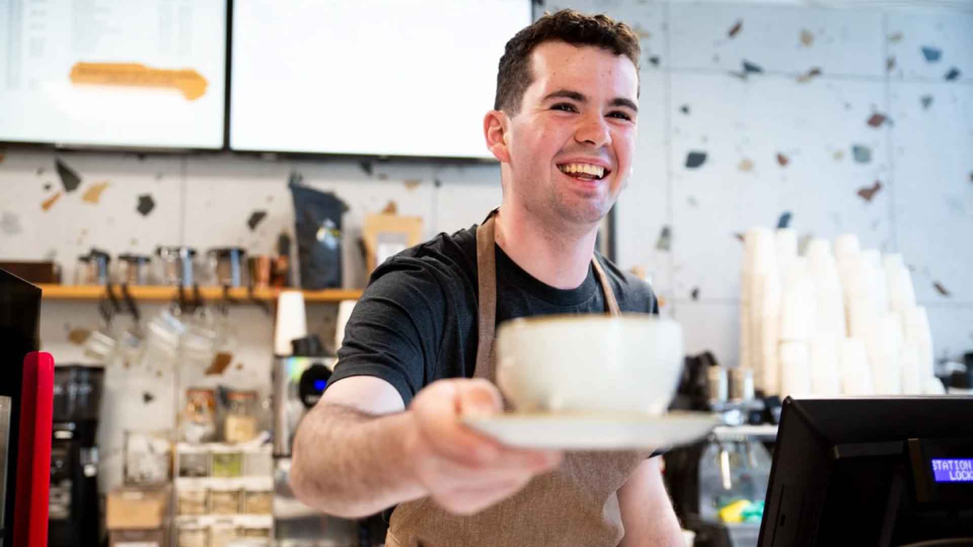 Student staff at Roasters cafe, smiling and handing someone a coffee from behind counter.