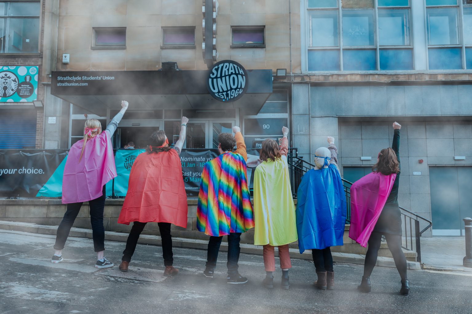 Students stood outside the Strath Union building wearing colourful capes and holding their fists in the air