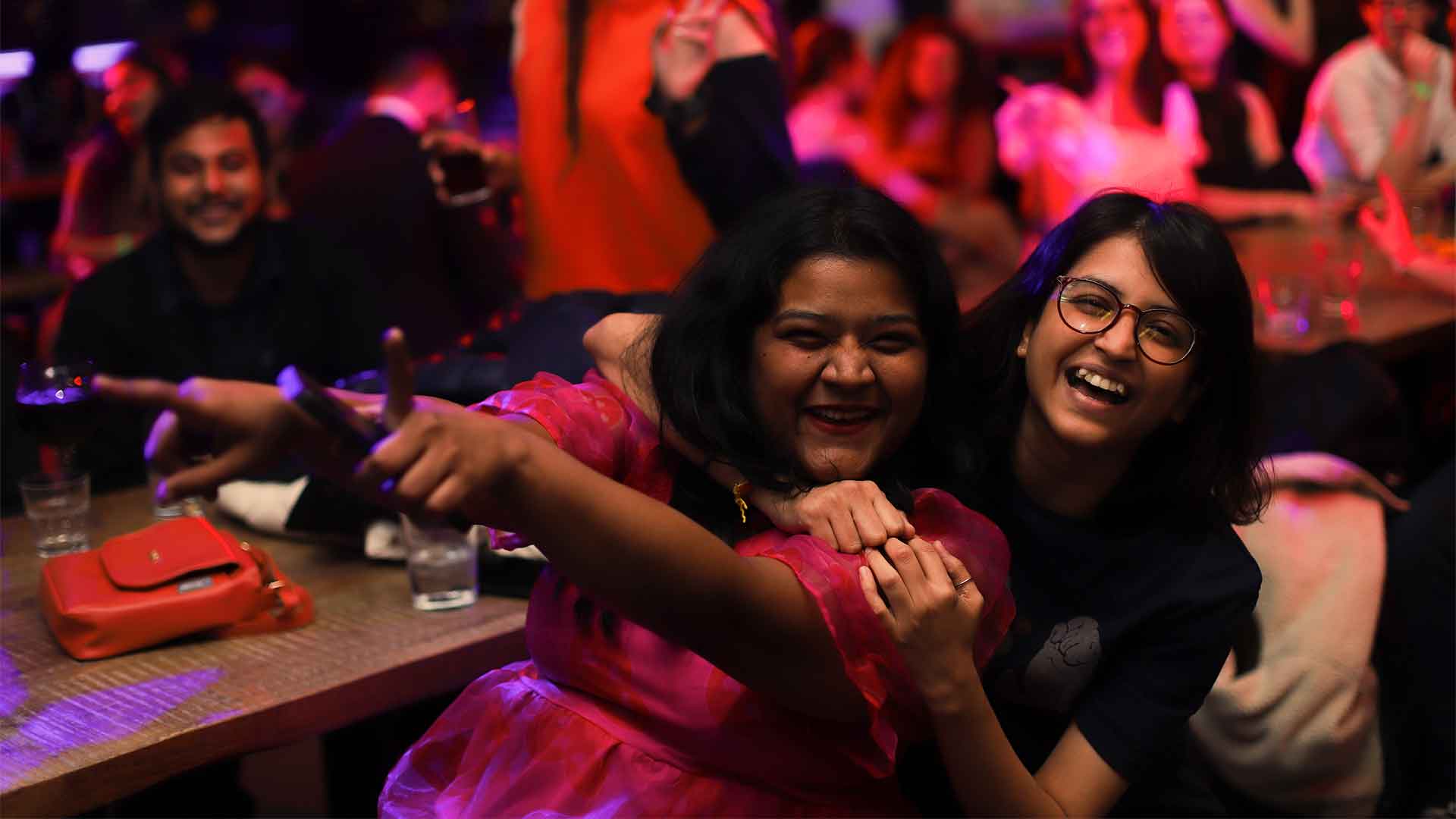 Two students sat at a wooden table laughing at the camera with their arms around each other
