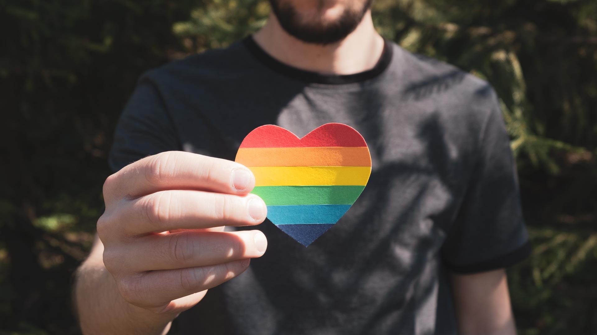 Person holding out a paper heart in the colours of the Pride flag.
