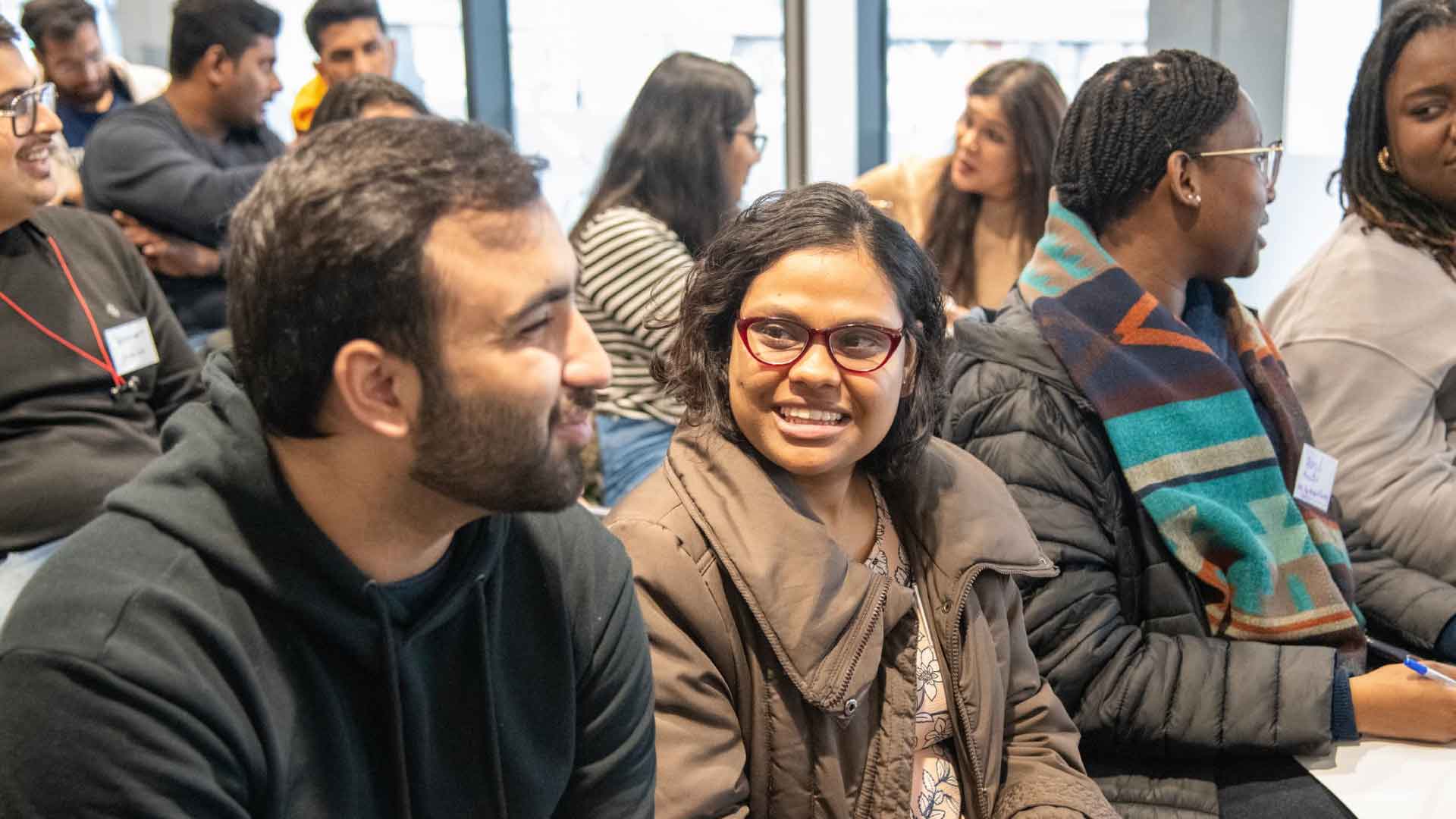 Two people smiling and discussing, with more people chatting in the background.
