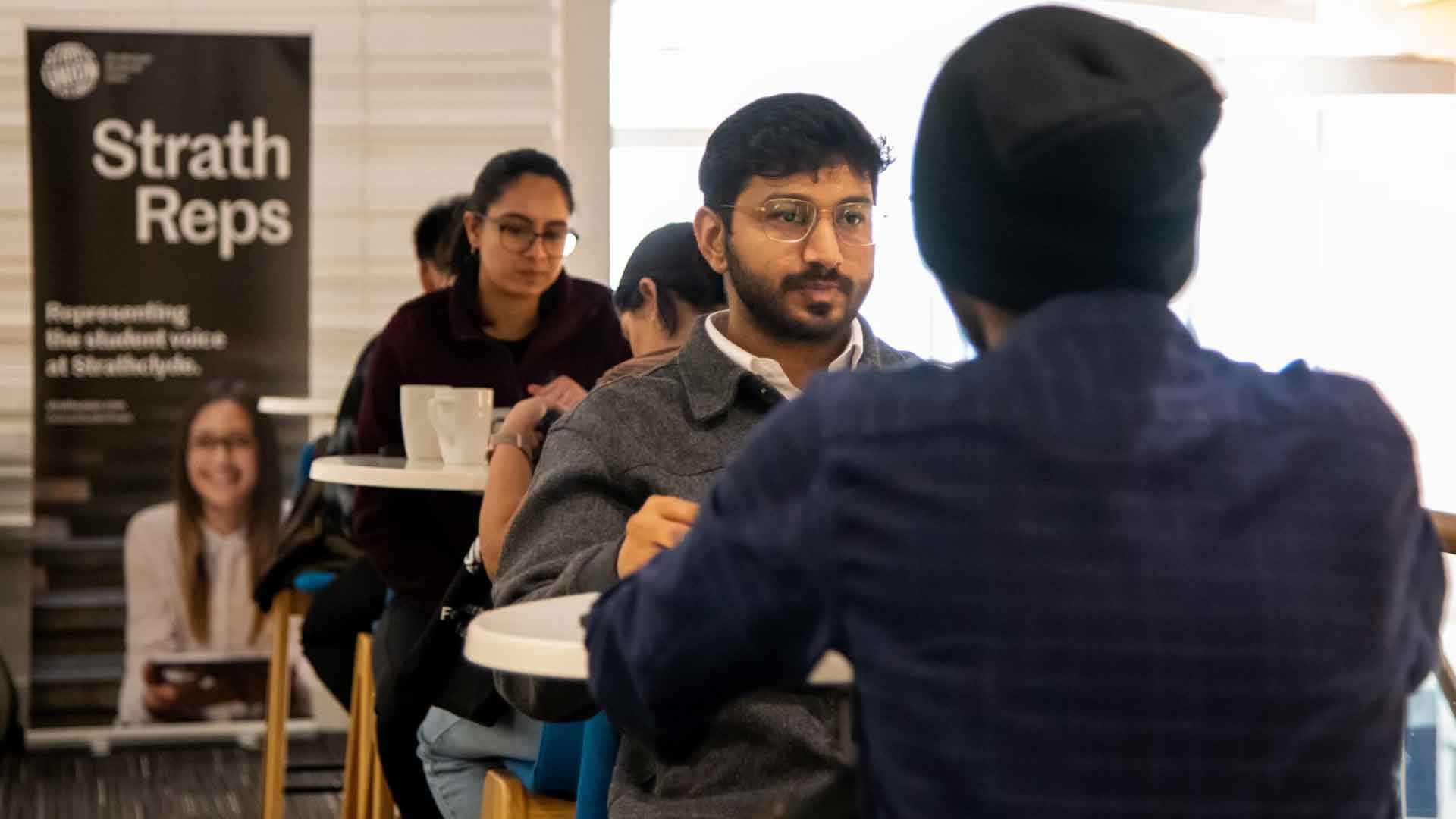 Students sitting around tables, discussing, with a 'Strath Reps' banner in the background.