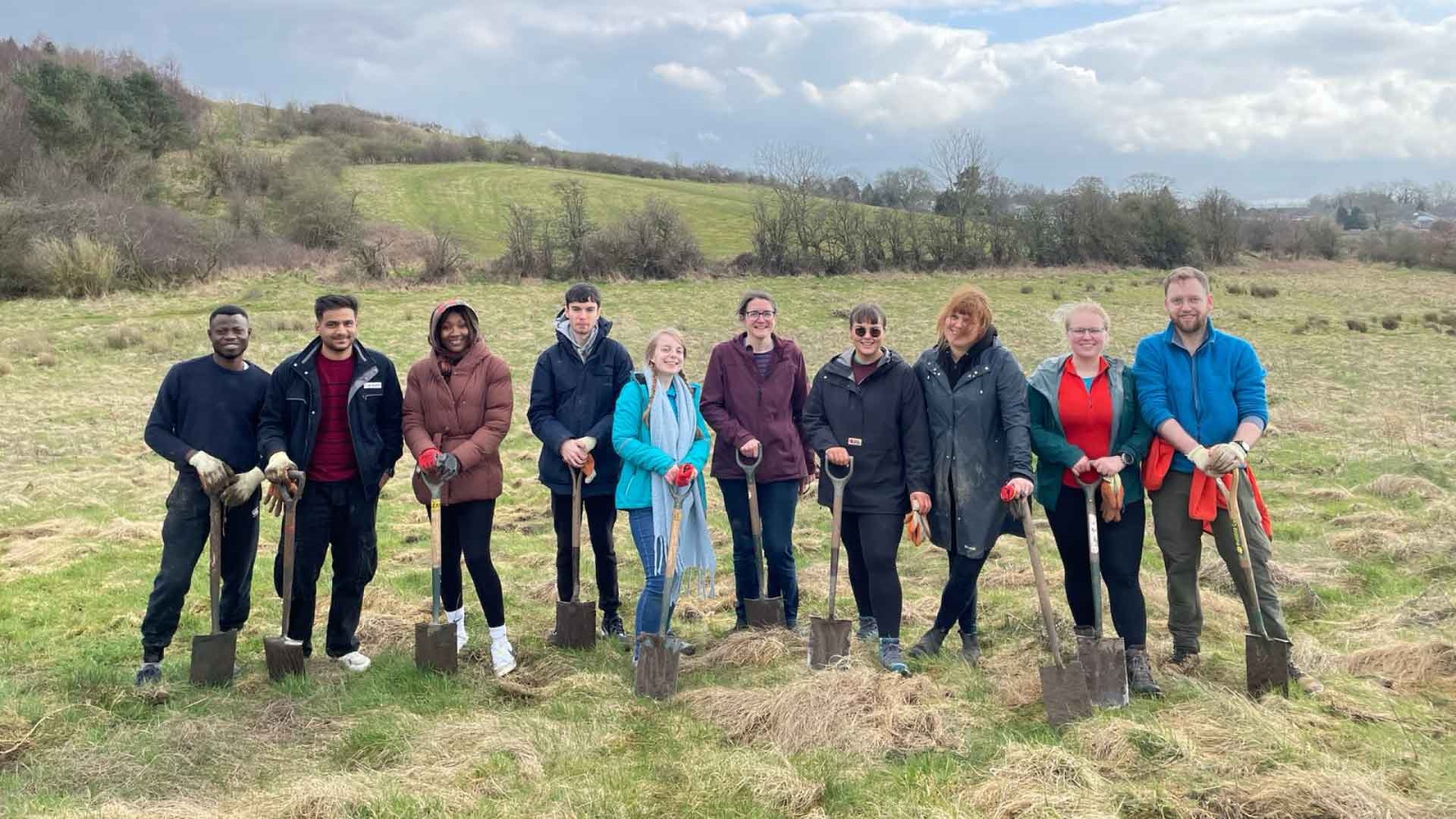 Strathclyde students and staff in the countryside, holding tree planting equipment and smiling at the camera.