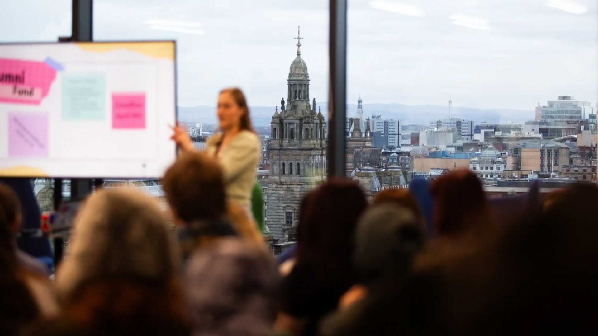 Person pointing to a screen, giving a speech to a crowd of students, with Glasgow visible in the background through a window.